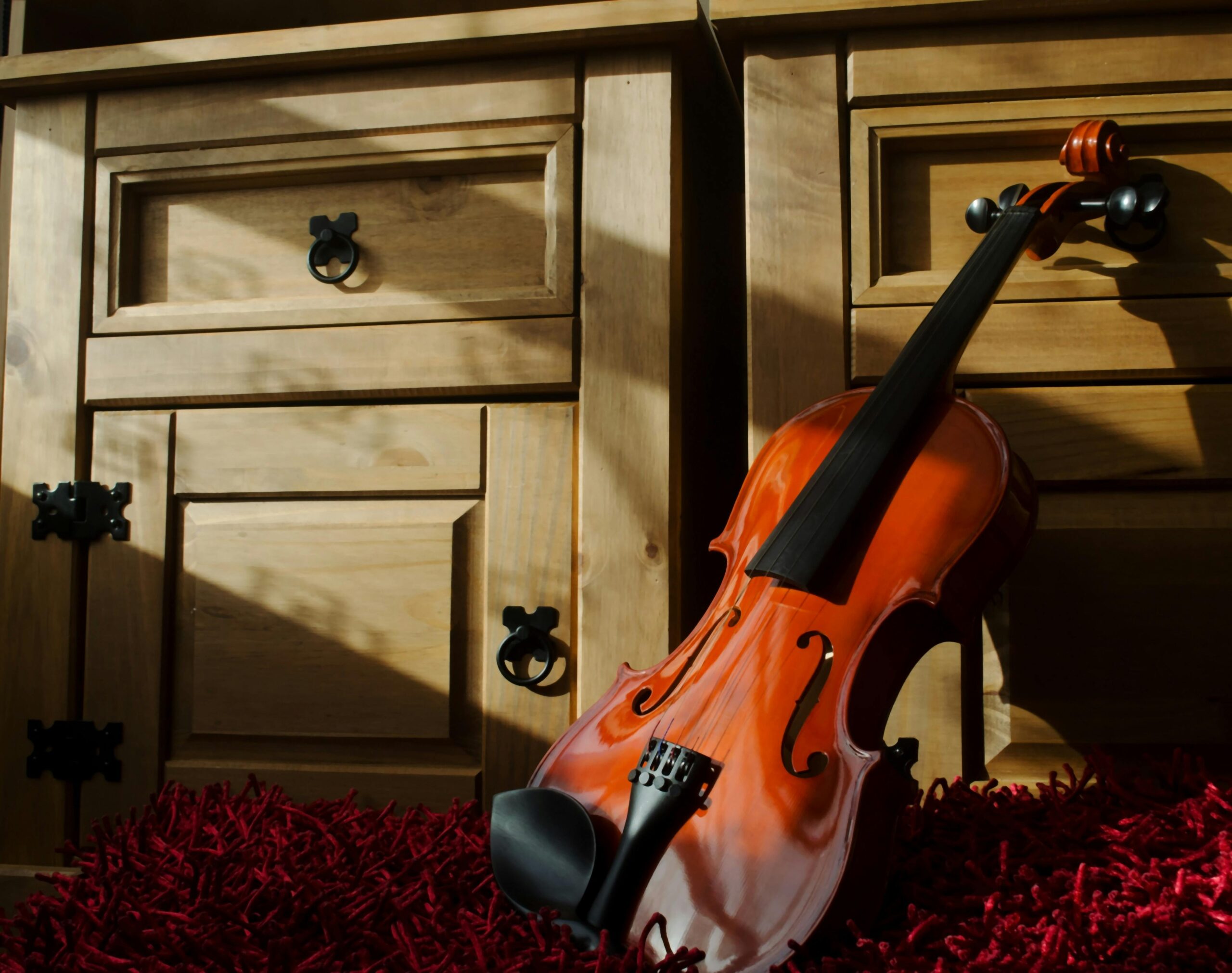 A beautifully crafted violin rests on a textured red mat, casting dramatic shadows against wooden drawers.